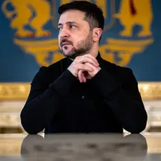 Prezydent Ukrainy Wołodymyr Zełenski w czarnej koszuli, z dłońmi złożonymi na stole, na tle flagi Ukrainy i herbu.