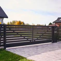 Nowoczesna, ciemnoszara brama przesuwna z poziomymi lamelami, otwierająca wjazd na posesję z kostką brukową.
