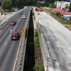 Widok z góry na wiadukt w budowie, z ruchem samochodowym po jednej stronie i placem budowy po drugiej.