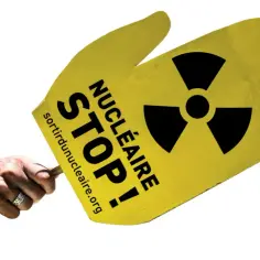Dłoń trzyma żółty znak z symbolem promieniowania i napisem "STOP NUCLEAIRE". Protest przeciwko energii jądrowej, która emituje niebezpieczne promieniowanie, w tym promieniowanie beta.
