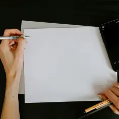 Dłoń trzymająca długopis nad kartką papieru, obok leży smartfon.