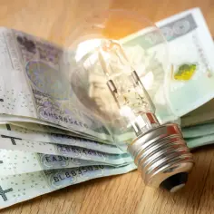 Żarówka na stosie polskich banknotów symbolizuje wysokie ceny energii. Bon energetyczny dla kogo? Dla każdego, kto zmaga się z rachunkami.