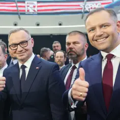 Dwóch mężczyzn w garniturach pokazuje kciuki w górę. Jeden z nich, w okularach, uśmiecha się. W tle widać baner z napisem "CPAC POLAND". Długość kadencji prezydenta jest tematem dyskusji.