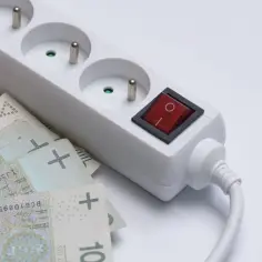 Biała listwa zasilająca z czerwonym włącznikiem i polskimi banknotami. To symboliczny dodatek do prądu, który pokazuje, jak drogi jest.