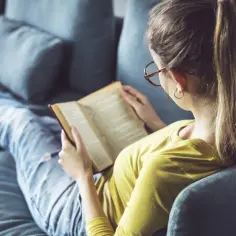 Dziewczyna w okularach relaksuje się na kanapie z książką. To jej ulubione lektury z gwiazdką, które pochłaniają ją bez reszty.