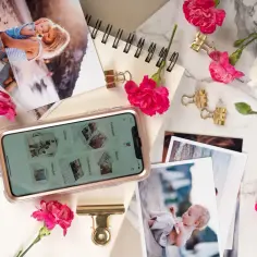 Telefon z aplikacją do wywoływania zdjęć, obok piękne fotografie rodzinne, kwiaty i złote spinacze.