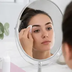 Kobieta w lustrze delikatnie zmywa hennę z brwi wacikiem. Szybka instrukcja, jak zmyć hennę z brwi.