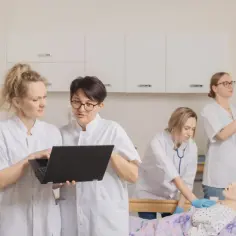 Pielęgniarki omawiają dane na laptopie, podczas gdy koleżanka ćwiczy zakładanie kroplówki. Zastanawiasz się, ile zarabia pielęgniarka po studiach magisterskich?