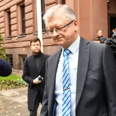 Mężczyzna w garniturze i krawacie, otoczony przez dziennikarzy z kamerami.