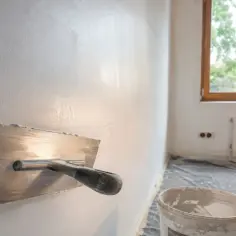 Paca do tynkowania zbliża się do świeżo nałożonej masy na ścianie. W tle okno i wiadro z zaprawą.