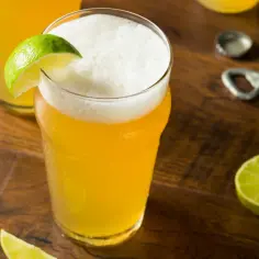 Jak zrobić domowy radler z piwa jasnego i lemoniady? Orzeźwiający napój z pianką i limonką na drewnianym blacie.