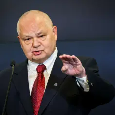 Prezes NBP w garniturze i czerwonym krawacie przemawia przy mikrofonie, gestykulując prawą ręką.