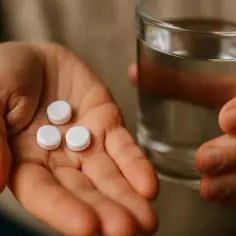 Dłoń trzyma trzy białe tabletki, prawdopodobnie **Panadol**, obok szklanka wody.