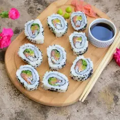 Pyszne california sushi z łososiem i awokado, podane z sosem sojowym i wasabi na drewnianej desce, ozdobione różowymi kwiatami.