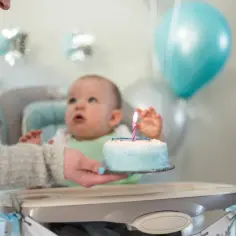Ciocia podaje tort na roczek. Dziecko wpatrzone w świeczkę, balony i ozdoby tworzą radosną atmosferę.
