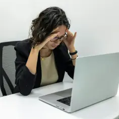 Kobieta z okularami, z rękami na skroniach, przy laptopie. Czy to objawy wypalenia zawodowego u kluczowego pracownika?