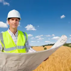 Inżynier w kasku i kamizelce odblaskowej analizuje plany, planując przekształcenie działki rolnej na budowlaną.
