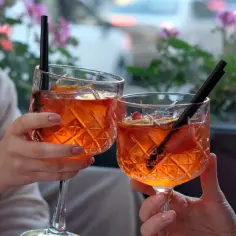 Dwie osoby wznoszą toast Aperolem bez alkoholu, ozdobionym owocami i lodem, na tle kwitnących kwiatów.