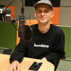 Vito Bambino w czarnej bluzie z napisem "bambino" siedzi przy stole w studiu nagraniowym.