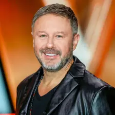Uśmiechnięty Andrzej Piaseczny, w czarnej skórzanej kurtce i z naszyjnikiem z koralików. Jego wiek dodaje mu charyzmy.