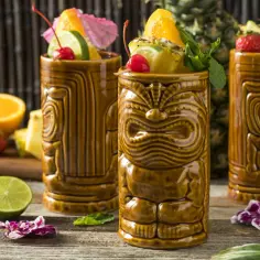 Trzy drinki tiki w brązowych kubkach z owocami i miętą. W tle ananas i cytrusy.