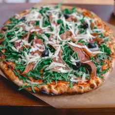 Pizza z rukolą, prosciutto, oliwkami i parmezanem na brązowym papierze.