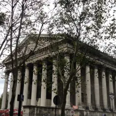 Majestatyczny kościół de la Madeleine w Paryżu, otoczony drzewami, z kolumnami w stylu korynckim.