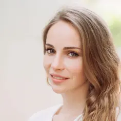 Uśmiechnięta kobieta z mysim kolorem włosów, delikatnie falowanymi, na jasnym, rozmytym tle.