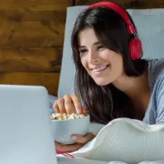 Uśmiechnięta dziewczyna w słuchawkach ogląda coś na laptopie, jedząc popcorn. To jej ulubiony sposób na relaks z multimedialnym odtwarzaczem.