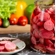 Słoik pełen różowych, soczystych kiszonych rzodkiewek. Na talerzyku kilka przekrojonych rzodkiewek. W tle warzywa: sałata, pomidory, ogórki i papryka.