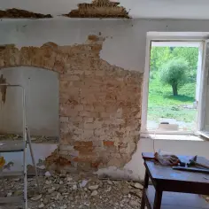 Remont w poniemieckich domach. Odkryta cegła, gruz i drabina wskazują na prace budowlane. Za oknem zieleń.