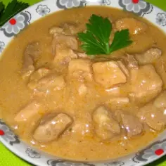 Gulasz z łopatki wieprzowej w kremowym sosie, udekorowany natką pietruszki, w misce z wzorem kwiatowym.