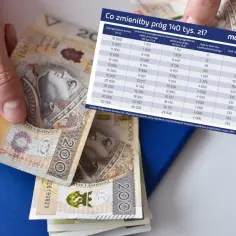Dłoń trzyma polskie banknoty 200 zł obok tabeli z danymi finansowymi.