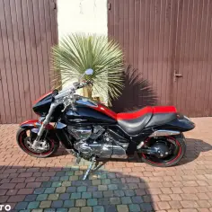 Czarny motocykl Suzuki Boulevard z czerwonym siedzeniem i detalami na felgach, zaparkowany na brukowanym podjeździe przed brązową bramą garażową.