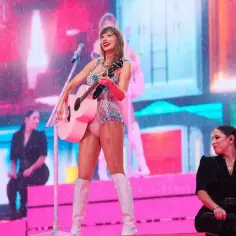 Taylor Swift, której wzrost jest imponujący, w błyszczącym stroju i białych kozakach gra na różowej gitarze.