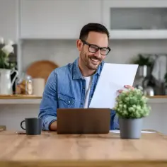Uśmiechnięty mężczyzna w okularach przegląda dokumenty przy laptopie w kuchni. To pokazuje, co to są media w mieszkaniu – praca i życie codzienne w jednym miejscu.