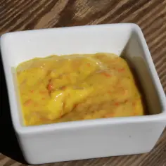 Gęsty, żółty dip na grilla z kawałkami marchewki w białej miseczce na drewnianym tle.