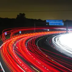 Nocna autostrada z czerwonymi i białymi smugami świateł samochodowych. Widać znaki drogowe, w tym informujące o minimalnej prędkości na autostradzie.