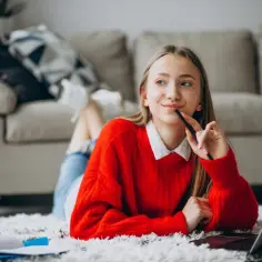 Dziewczyna w czerwonym swetrze uczy się w domu, korzystając z laptopa. To jej nauczanie domowe w liceum.
