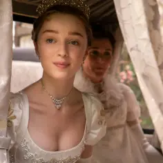 Phoebe Dynevor w stroju z epoki, z tiarą i naszyjnikiem, w karocy. Jej filmy, seriale i programy to często historie kostiumowe.