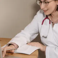 Lekarka w białym kitlu z czerwonym stetoskopem analizuje dane na laptopie i notuje w zeszycie. Dowiaduje się, jak zostać lekarzem medycyny pracy.