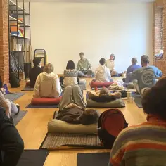 Grupa osób na zajęciach, ucząca się, czym jest mindfulness. Siedzą na poduszkach, w ciszy i spokoju.
