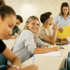 Uśmiechnięta studentka pisze notatki, zastanawiając się, co po geografii rozszerzonej. W tle nauczycielka z żółtym folderem.