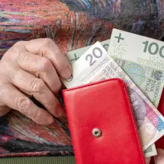 Dłoń starszej osoby wkłada pieniądze do czerwonego portfela. Widać banknoty 20 zł i 100 zł. Zastanawiasz się, ile wynosi dodatek mieszkaniowy?