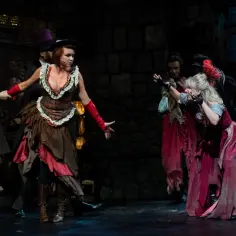 Nędznicy teatr muzyczny: kobiety w strojach z epoki, w tym jedna w brązowo-czerwonej sukni, tańczą na scenie.