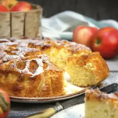 Wilgotne ciasto z jabłkami na oleju, posypane cukrem pudrem, obok świeże jabłka i kawałek ciasta na talerzyku.