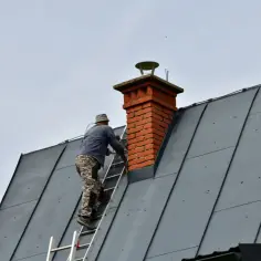 Kominiarz wspina się po drabinie do ceglanego komina. Przypomnienie o tym, jak często czyścić komin, przepisy są ważne dla bezpieczeństwa.