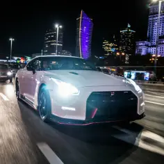 Biały Nissan GT-R Nismo mknie nocą przez miasto, oświetlony reflektorami i neonami wieżowców.