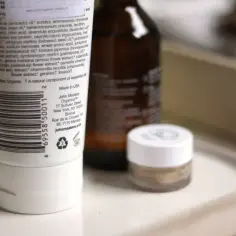 Biała tubka z kosmetykiem John Masters Organics, z widoczną listą składników, w tym ethylhexylglycerin. Obok butelka i mały słoiczek.