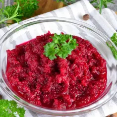 Ćwikła z chrzanem w szklanej misce, ozdobiona natką pietruszki, na tle serwetki i ziół.